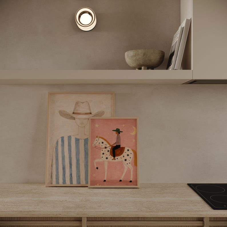 May include: Two framed art prints on a countertop. One print features a person in a cowboy hat with blue and white stripes. The other print shows a person riding a horse with star patterns on a pink background. A small bowl and a light fixture are also visible.