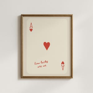 Puede incluir: Impresión artística enmarcada con el diseño de la carta As de Corazones. La tarjeta tiene un fondo crema con corazones rojos y el texto "how lucky are we". El marco es dorado.