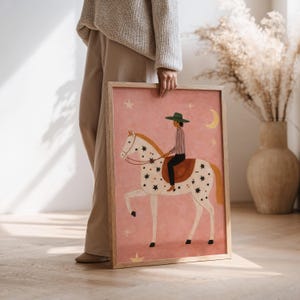 May include: A framed art print featuring a whimsical illustration of a rider on a white horse adorned with star patterns. The artwork has a pink background with celestial elements and a wooden frame.