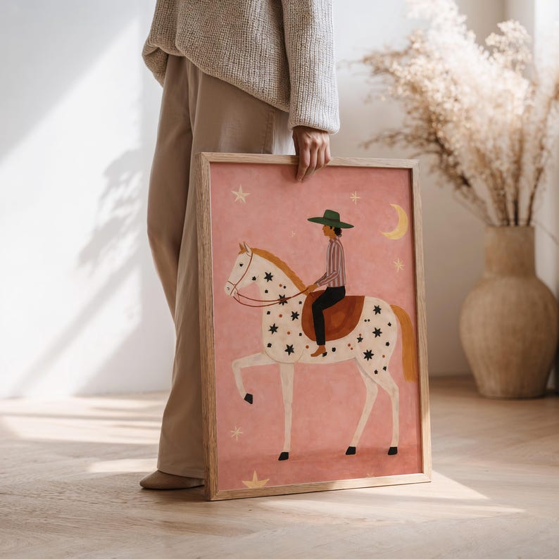 May include: A framed art print featuring a whimsical illustration of a rider on a white horse adorned with star patterns. The artwork has a pink background with celestial elements and a wooden frame.