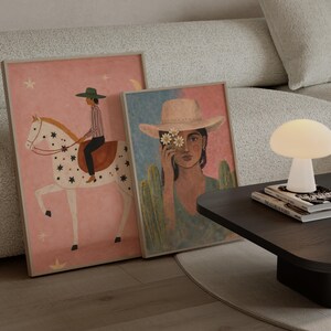 May include: Two framed art prints lean against a sofa. One print features a person riding a white horse with star details on a pink background. The other print depicts a person wearing a hat with flowers, set against a blue and green backdrop. A mushroom-shaped lamp sits on a dark table.