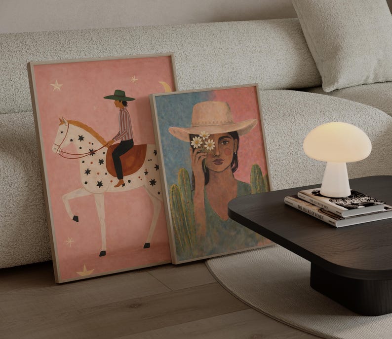 May include: Two framed art prints lean against a sofa. One print features a person riding a white horse with star details on a pink background. The other print depicts a person wearing a hat with flowers, set against a blue and green backdrop. A mushroom-shaped lamp sits on a dark table.