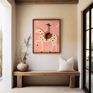 May include: A framed art print featuring a person riding a horse with a star and moon design on a pink background. The artwork is in a wooden frame and hangs above a wooden bench with a white pillow.