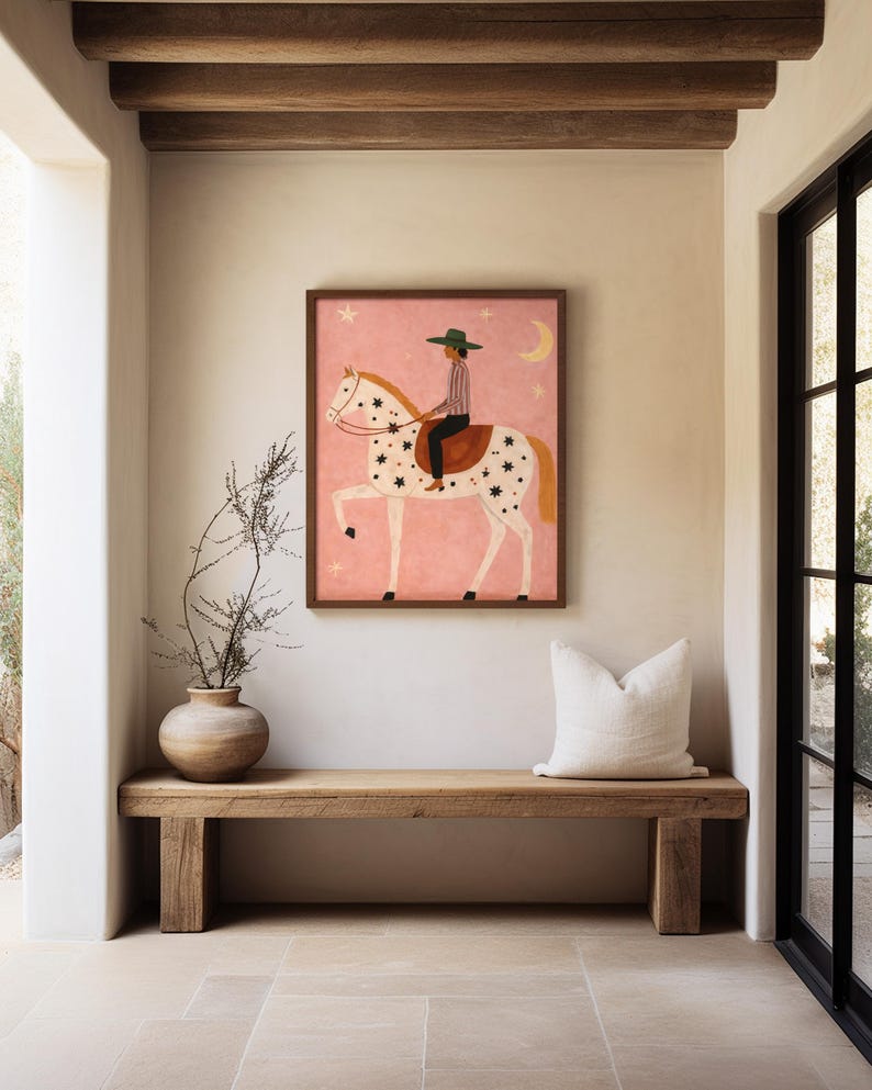 May include: A framed art print featuring a person riding a horse with a star and moon design on a pink background. The artwork is in a wooden frame and hangs above a wooden bench with a white pillow.