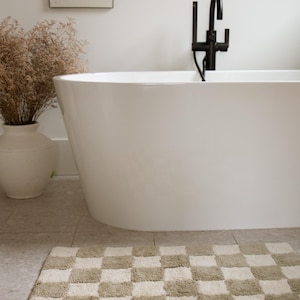 May include: A white, oval-shaped bathtub with a black faucet is positioned against a white wall. A checkered bath mat in beige and cream tones sits on the floor. A vase with dried plants and a framed artwork complete the bathroom scene.