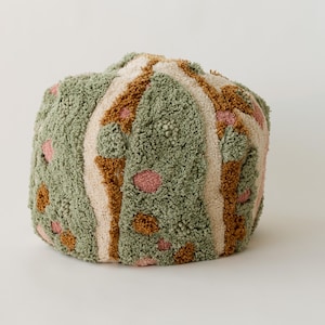 Op de afbeelding: Een ronde, getextureerde poef in tinten groen, beige, bruin en roze. De poef heeft een getuft ontwerp met verticale beige strepen en verspreide roze en bruine cirkelvormige accenten. De algehele esthetiek is zacht en uitnodigend.