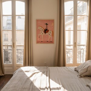 May include: A framed art print featuring a whimsical illustration of a person riding a spotted horse under a crescent moon and stars. The artwork is set against a pink background and hangs on a white wall. The room has large windows and neutral-colored curtains.
