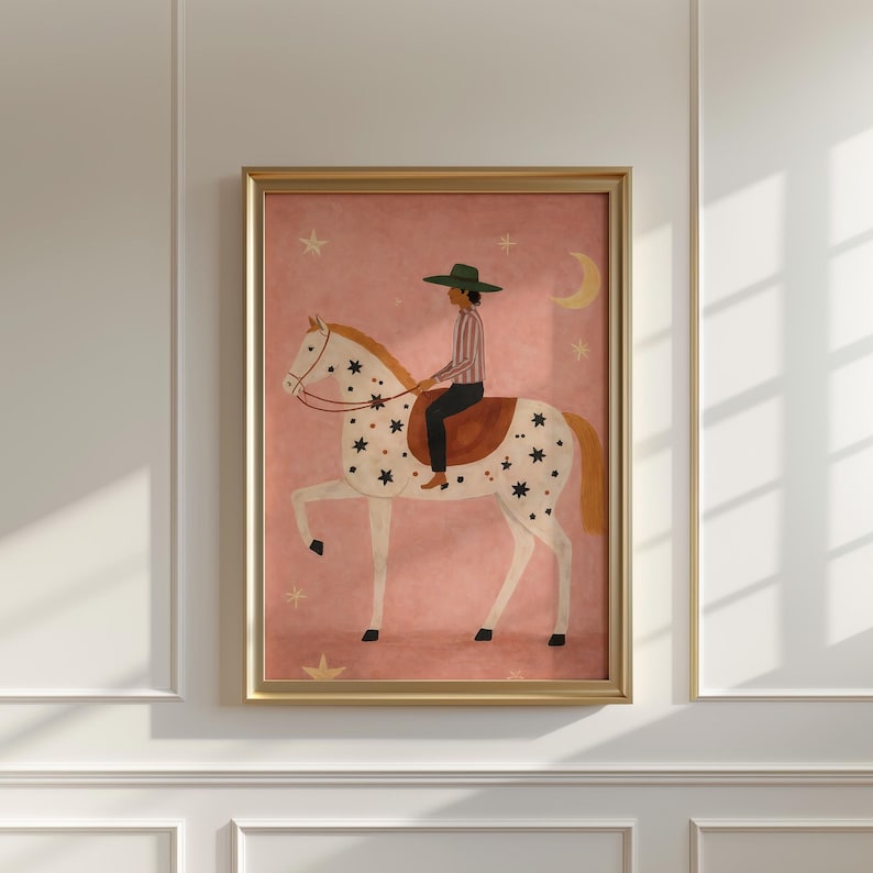 May include: A framed art print featuring a whimsical illustration of a person riding a horse. The horse is white with black star-shaped markings. The rider wears a green hat and striped shirt. The background is pink with stars and a crescent moon.