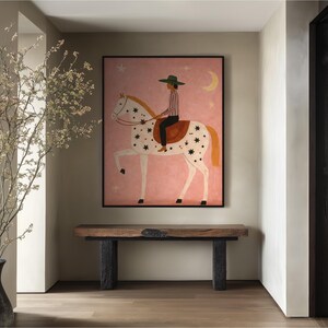 May include: A framed artwork depicts a person riding a white horse with black star-shaped markings against a pink background. The rider wears a green hat and striped shirt. A wooden bench with black legs sits below the artwork. A vase with branches is on the left.