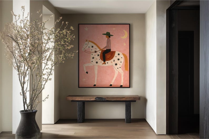 May include: A framed artwork depicts a person riding a white horse with black star-shaped markings against a pink background. The rider wears a green hat and striped shirt. A wooden bench with black legs sits below the artwork. A vase with branches is on the left.