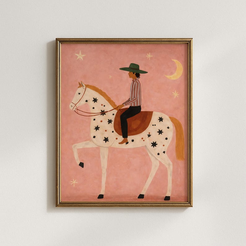 May include: A framed art print featuring a whimsical illustration of a rider on a white horse adorned with black stars. The rider wears a green hat and striped shirt. The background is a soft pink with stars and a crescent moon.