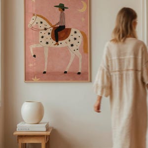 May include: A framed art print depicts a person riding a horse adorned with stars against a pink background. The rider wears a green hat and striped shirt. A crescent moon and stars are also in the background. A white vase and books sit on a wooden table.