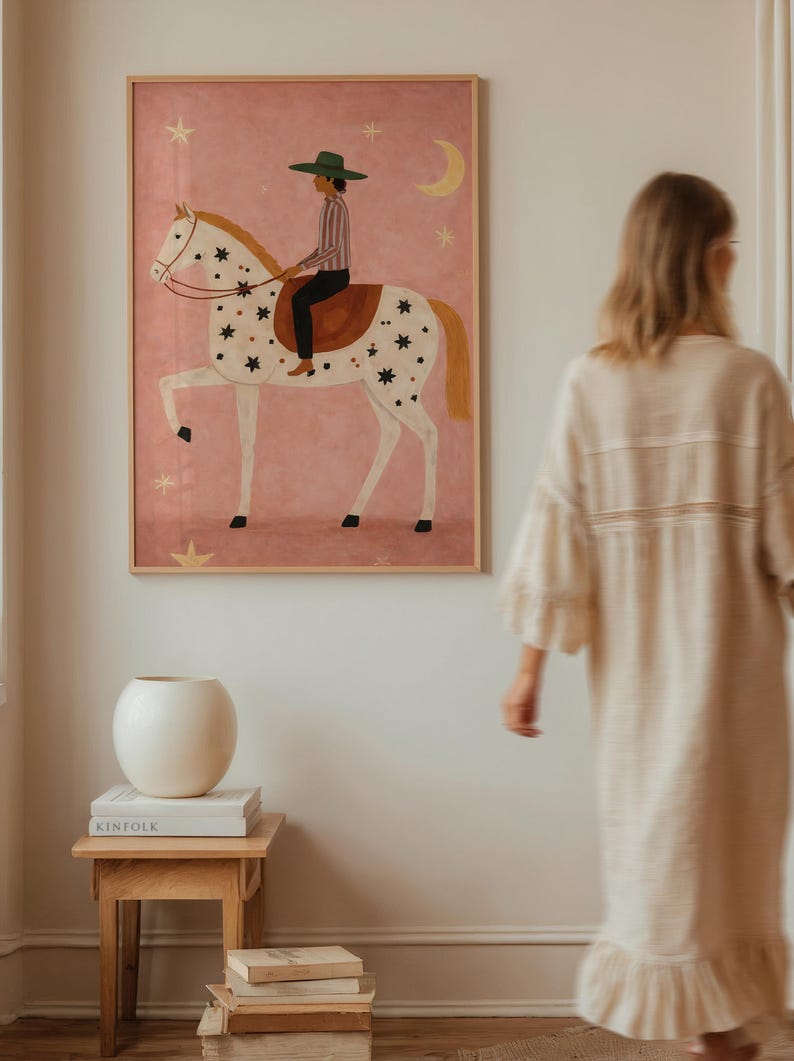 May include: A framed art print depicts a person riding a horse adorned with stars against a pink background. The rider wears a green hat and striped shirt. A crescent moon and stars are also in the background. A white vase and books sit on a wooden table.