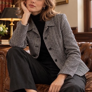 May include: A black and white herringbone patterned blazer with a collared neckline and a front button closure. The blazer is worn over a solid black mock neck top and paired with dark gray trousers, creating a structured and layered look.