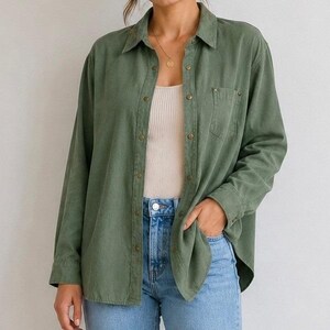 Può includere: Una camicia a maniche lunghe verde oliva con abbottonatura, colletto, una tasca sul petto e bottoni metallici. La camicia è indossata aperta sopra una canotta bianca a coste, abbinata a jeans in denim azzurro con bottone metallico visibile.