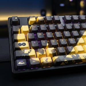 Puede incluir: Un teclado mecánico negro y amarillo con teclas personalizadas. Las teclas presentan un diseño de noche estrellada e ilustraciones de personajes. El teclado tiene una distribución estándar con teclas de función y una tecla de control.