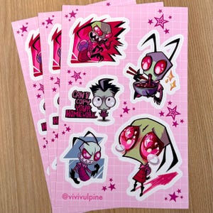 May include: Pink sticker sheets featuring cartoon alien characters in various poses. One sticker reads "Can I Copy Your Homework?" with a character. The stickers have a pink background with star accents. The artist's handle is @vivivulpine.