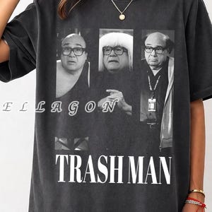 May include: Dark gray t-shirt featuring a black and white image of a person in three different poses. The word "TRASH MAN" is printed in white block letters below the image. The brand name "NELLAGON" is printed in a cursive font.