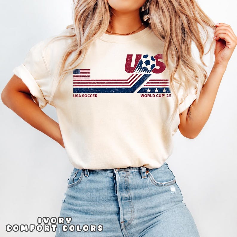 May include: An ivory short-sleeve t-shirt featuring a graphic design with the text USA SOCCER WORLD CUP 26. The design includes a soccer ball, red and blue horizontal stripes, stars, and a stylized American flag motif in a distressed texture.
