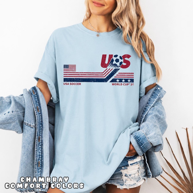 May include: A light blue t-shirt featuring a graphic design with the text USA SOCCER WORLD CUP '26. The design includes a stylized soccer ball, red and blue horizontal stripes, and a small American flag. The shirt is worn with a denim jacket.
