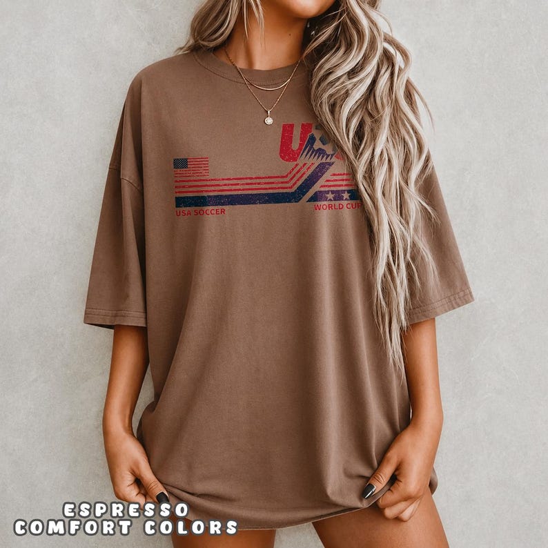 May include: A brown short-sleeve t-shirt featuring a distressed graphic print with red and blue horizontal stripes, stars, and the text USA SOCCER WORLD CUP. The design includes a stylized soccer ball icon and a small American flag graphic element.