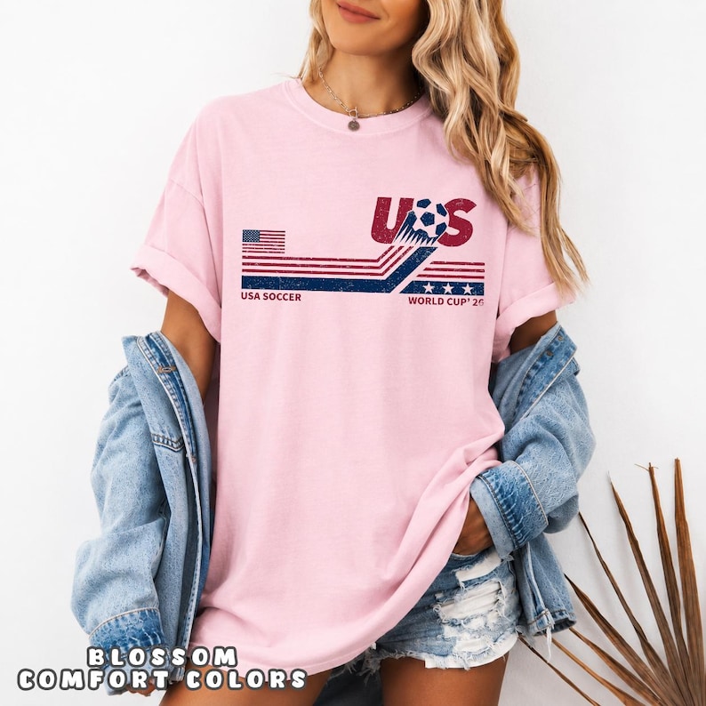 May include: A light pink short-sleeve t-shirt featuring a graphic design with the text USA SOCCER and WORLD CUP '26. The design includes a stylized soccer ball, red and blue horizontal stripes, a small American flag, and three blue stars.