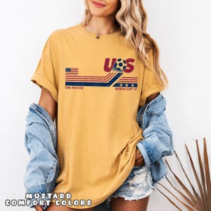 May include: A mustard yellow short-sleeve t-shirt featuring a distressed graphic with the text USA SOCCER WORLD CUP 2026. The design includes a stylized soccer ball, red and blue horizontal stripes, a small American flag, and three stars.