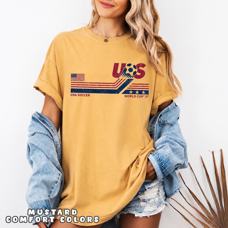 May include: A mustard yellow short-sleeve t-shirt featuring a distressed graphic with the text USA SOCCER WORLD CUP 2026. The design includes a stylized soccer ball, red and blue horizontal stripes, a small American flag, and three stars.