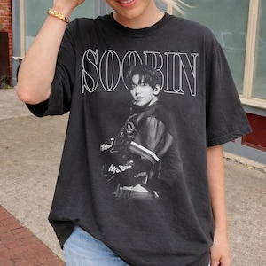 May include: Black graphic t-shirt featuring a black and white image of a person wearing a jacket. The word "SOOBIN" is printed in white above the image. The shirt is a dark gray color.