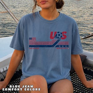 May include: A blue jean colored t-shirt featuring a distressed graphic design with the text USA and WORLD CUP 26. The print includes a soccer ball, red and blue horizontal stripes, a small American flag, and three stars in a weathered style.
