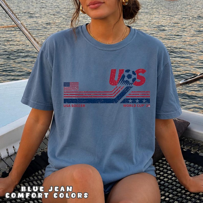 May include: A blue jean colored t-shirt featuring a distressed graphic design with the text USA and WORLD CUP 26. The print includes a soccer ball, red and blue horizontal stripes, a small American flag, and three stars in a weathered style.