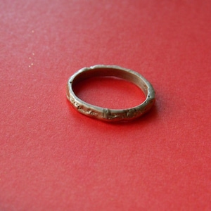 May include: A gold-colored ring with a textured surface. The ring is set against a red background. The ring's design appears to be slightly uneven, with a rustic or antique aesthetic.