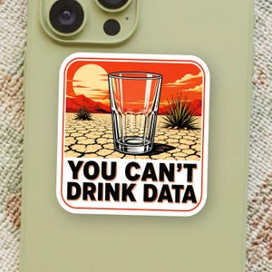 May include: A square sticker featuring a desert landscape with a sunset, a glass in the center, and the text YOU CAN'T DRINK DATA in bold black letters. The background shows cracked earth, distant mountains, and spiky desert plants under an orange sky.