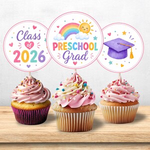 Peut inclure: Trois cupcakes surmontés de décorations en papier circulaires. La première indique Class of 2026 avec des cœurs. La deuxième affiche PRESCHOOL Grad avec un arc-en-ciel et un soleil. La troisième présente une toque violette. Glaçage rose.