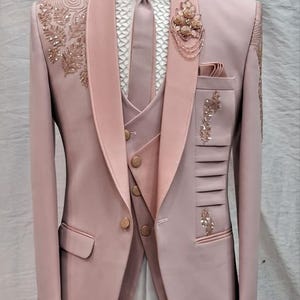 Könnte beinhalten: Ein formeller rosa Anzug mit Schalkragen, Weste und Krawatte, präsentiert auf einer Schaufensterpuppe. Der Anzug hat aufwendige Stickereien auf Revers und Tasche, mit goldfarbenen Knöpfen. Eine dekorative Brosche ziert das Revers. Ein formelles Kleidungsensemble.