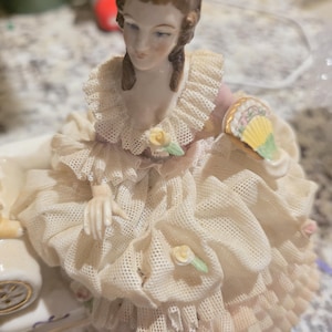 Peut inclure: Figurine en porcelaine d'une femme dans une robe crème avec des bordures en dentelle et un éventail. La figurine a un visage détaillé et est ornée de petites fleurs délicates. La robe a plusieurs couches de dentelle et de volants.