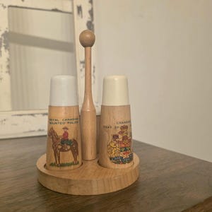 Könnte beinhalten: Salz- und Pfefferstreuer-Set aus Holz mit einem zentralen Holzgriff. Das Set enthält zwei zylindrische Streuer mit weißen Deckeln, die jeweils mit einem anderen Bild und Text im Zusammenhang mit der Royal Canadian Mounted Police und dem kanadischen Wappen verziert sind.