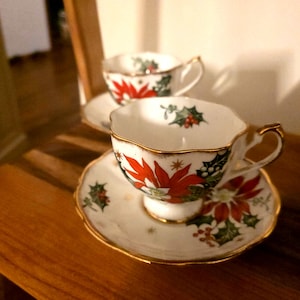 May include: Two white teacups and saucers with gold trim and a Christmas poinsettia and holly design. The teacups are on a wooden surface. The teacups have a gold handle and a gold trim around the rim.