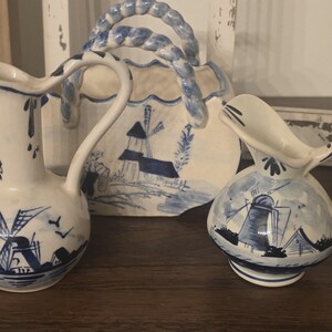 Può includere: Brocche e cestino in ceramica bianca e blu con disegni di mulini a vento. Le brocche hanno manici e beccucci curvi. Il cestino ha un manico intrecciato. I disegni sono in stile Delft.