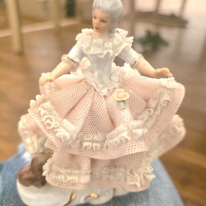 Könnte beinhalten: Porzellanfigur einer Frau in einem rosa-weißen Kleid mit aufwendigen Spitzendetails. Die Figur hat eine gepuderte Perücke und ist mit ausgestreckten Armen posiert. Der Sockel ist weiß mit goldenen Akzenten, im Rokoko-Stil.