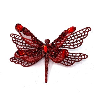 May include: A decorative dragonfly ornament in shades of red. The wings are a delicate openwork design with glitter accents and red gemstones. The body and head are also covered in glitter. The dragonfly is set against a white background.
