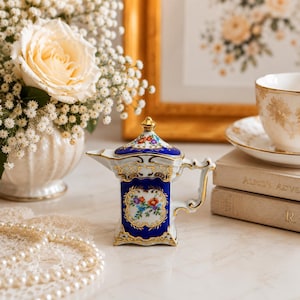 May include: A small porcelain creamer with a rectangular body, featuring a deep blue glaze, gold filigree accents, and a central panel decorated with a colorful floral bouquet. It has a matching lid with a gold finial and a curved handle.