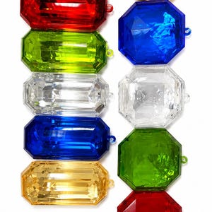 Puede incluir: Una colección de diez gemas de plástico facetadas dispuestas en dos columnas verticales. La columna izquierda presenta cinco gemas rectangulares en rojo, verde lima, transparente, azul y dorado. La columna derecha presenta cinco gemas octogonales en dorado, azul, transparente, verde y rojo.
