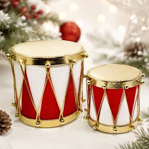May include: Two cylindrical toy drums featuring a white and red triangular pattern on the sides. Each drum has a metallic gold top and bottom rim, connected by thin gold tension rods with small gold spheres at the top and bottom edges.