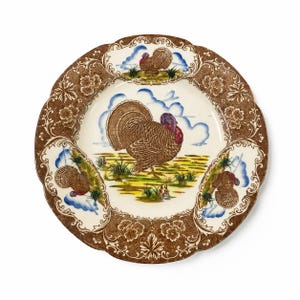 May include: A round ceramic dinner plate featuring a central illustration of a brown turkey in a field with blue clouds. The scalloped rim is decorated with a brown floral pattern and three smaller turkey motifs set against blue cloud accents.