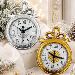 May include: Two round hanging ornaments shaped like clocks, one in silver and one in gold. Each features a white face with black Roman numerals, black clock hands, and a decorative bow at the top. The silver clock has a silver string, while the gold clock has a gold string for hanging.