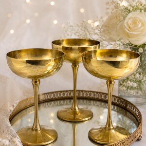 May include: Three polished brass coupe glasses with wide, shallow bowls and slender stems, arranged on a round, mirrored serving tray with a decorative gold-toned metal rim featuring an intricate, perforated lattice pattern.