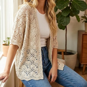 Könnte beinhalten: Eine beigefarbene, vorne offene Häkelstrickjacke mit einem detaillierten, muschelförmigen Spitzenmuster, kurzen Ärmeln und geradem Saum, getragen über einem schlichten weißen Tanktop und einer blauen Jeans.