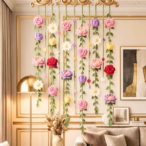 Peut inclure: Une décoration murale florale avec des fleurs crochetées roses, rouges, jaunes, violettes et blanches. Les fleurs sont attachées à des lianes vertes et suspendues à un élément décoratif doré.