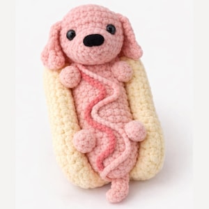 Puede incluir: Un animal de peluche tejido a ganchillo con forma de perro rosa metido dentro de un panecillo de color crema. El perro tiene ojos de plástico negro, una nariz negra y una línea de hilo rosa ondulada en el cuerpo que imita una salsa de hot dog.